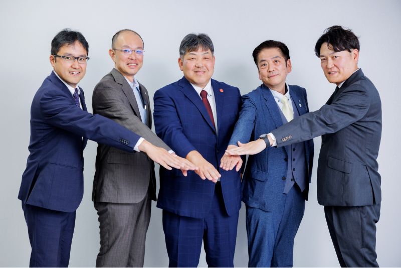 写真:令和7年度支援先企業の皆さんが笑顔で手を組むようす